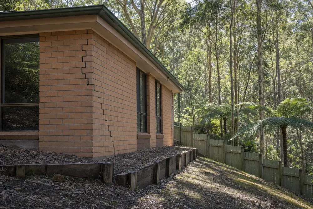 A brick house on a steep block with a stair-step crack at its corner, a timber-sleeper edge below it and forest behind — underpinning, Melbourne. Illustrative example.