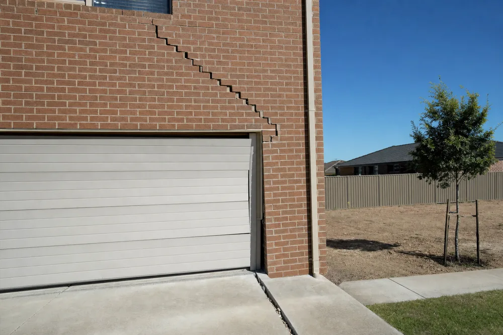 A brick house wall with a wide stair-step crack rising from the corner of a garage door, beside an open joint in the driveway — underpinning, Melbourne. Illustrative example.