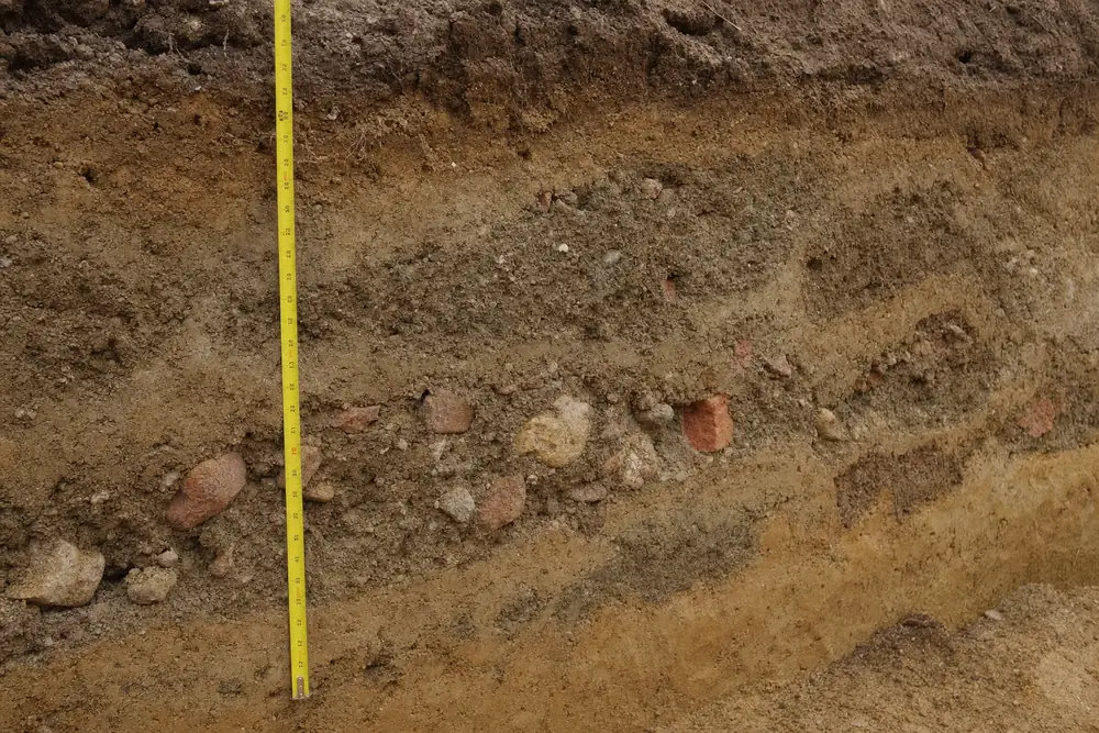 A trench wall showing soil with pockets of brick fragments and stones, a tape measure against it — underpinning, Melbourne. Illustrative example.
